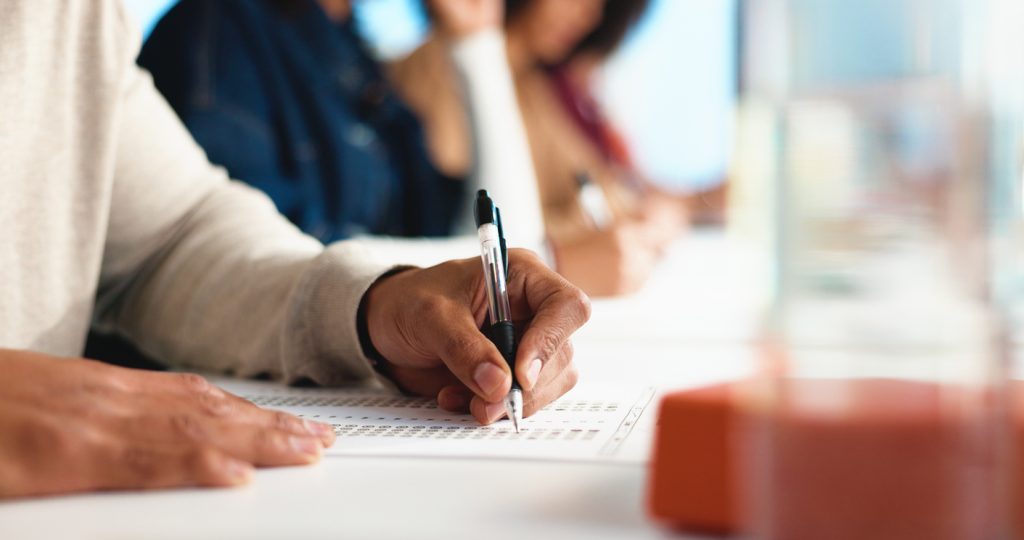 A group of people filling out forms next to each other. Their faces are not visible, and the people in the background are blurry.